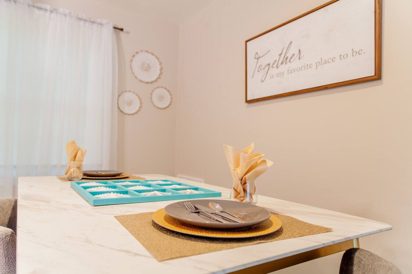 A dining table set with plates, cutlery, and napkins, with a framed quote on the wall.