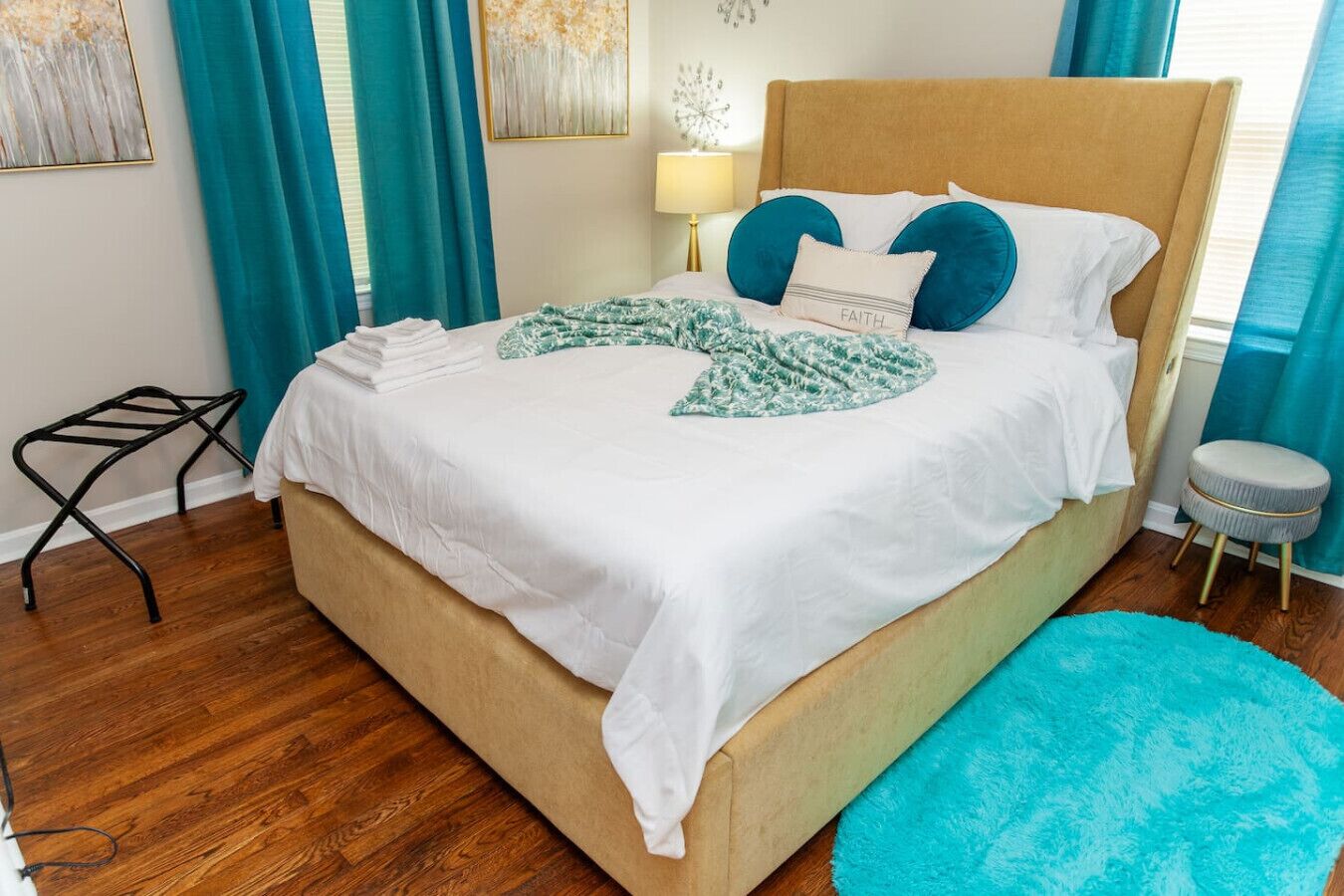 Una cama bien hecha con un edredón blanco y almohadas turquesa, un taburete pequeño y una alfombra azul en una habitación con suelo de madera y cortinas turquesa.