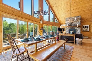 Une cabane en bois spacieuse, dotée de grandes fenêtres, d’un coin repas, d’une cheminée et d’un lit en mezzanine.