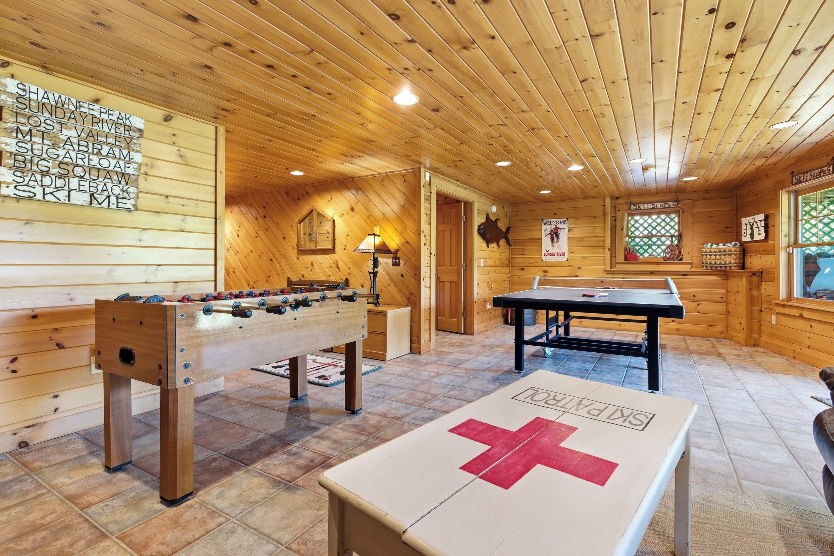 A wooden-floored room with a foosball table, a table tennis table, and a foosball table with a red cross.