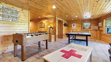 A wooden-floored room with a foosball table, a table tennis table, and a foosball table with a red cross.