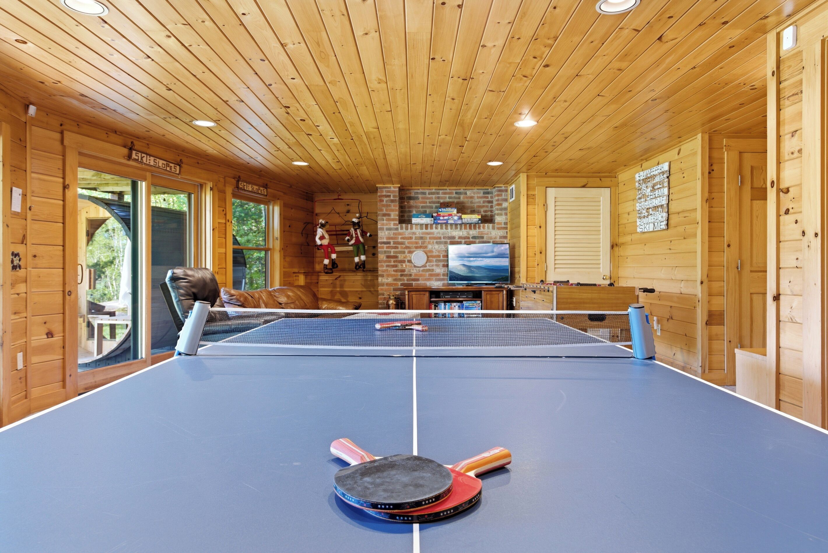 A ping pong table in a wooden cabin with a brick fireplace and a view of the outdoors.
