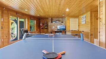 A ping pong table in a wooden cabin with a brick fireplace and a view of the outdoors.