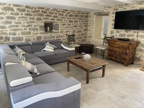 A living room with a stone wall, a grey sectional sofa, a wooden coffee table, a small table with a chair, and a TV mounted on the wall. - Stone house - Rocamadour 7 min - 10/12 Pers (Rignac)