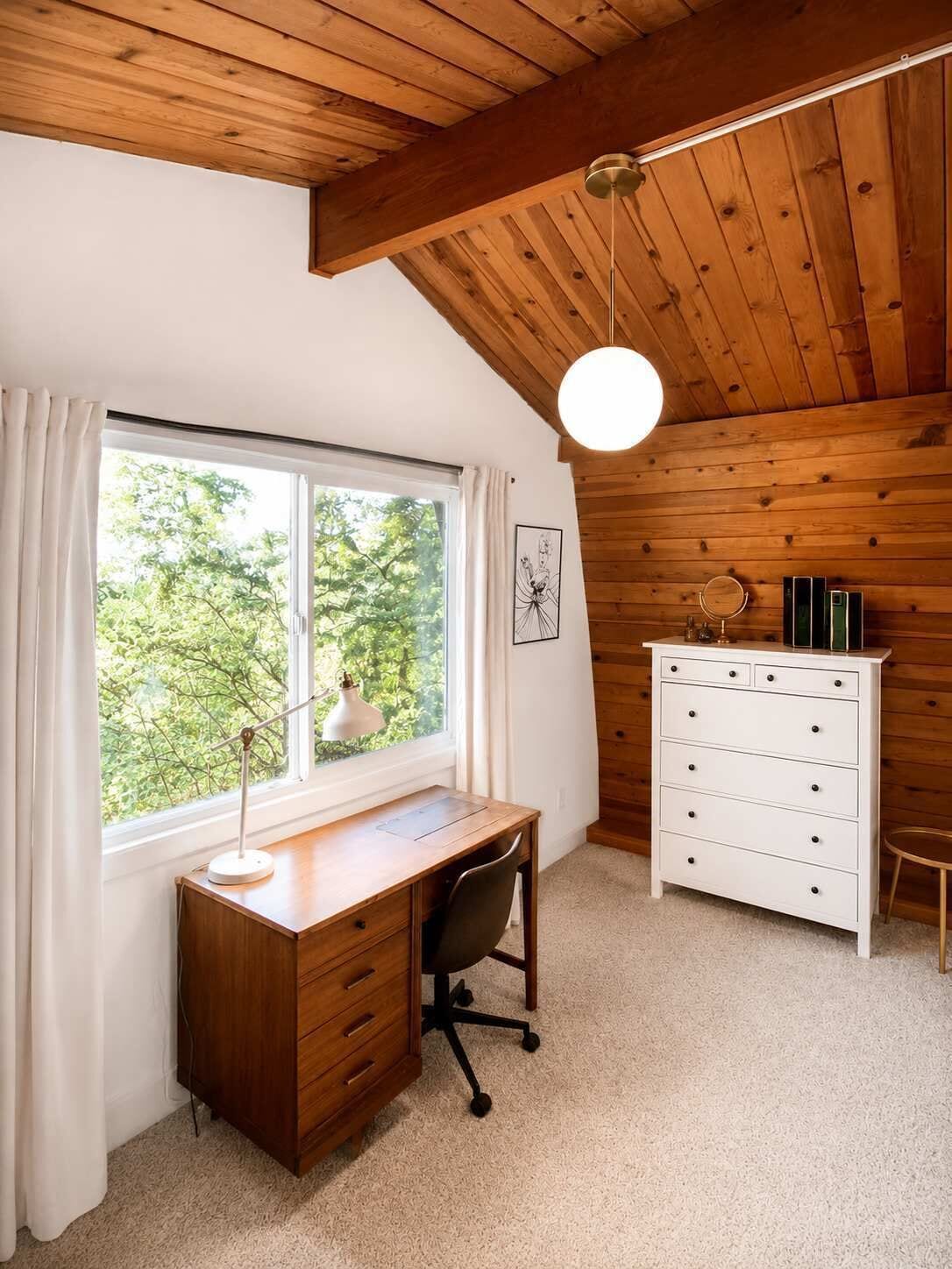 Una habitación con techo de madera, un escritorio con cajones, un aparador blanco y una ventana con cortinas.