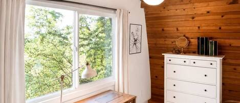 Una habitación con techo de madera, un escritorio con cajones, un aparador blanco y una ventana con cortinas.