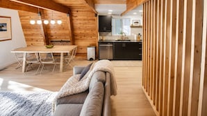 A cozy interior with a wooden ceiling, a dining area with a table and chairs, a kitchen with dark cabinets, and a gray sofa with a blanket. - Modern & Cozy, Family-Friendly Cabin (Lake Arrowhead)