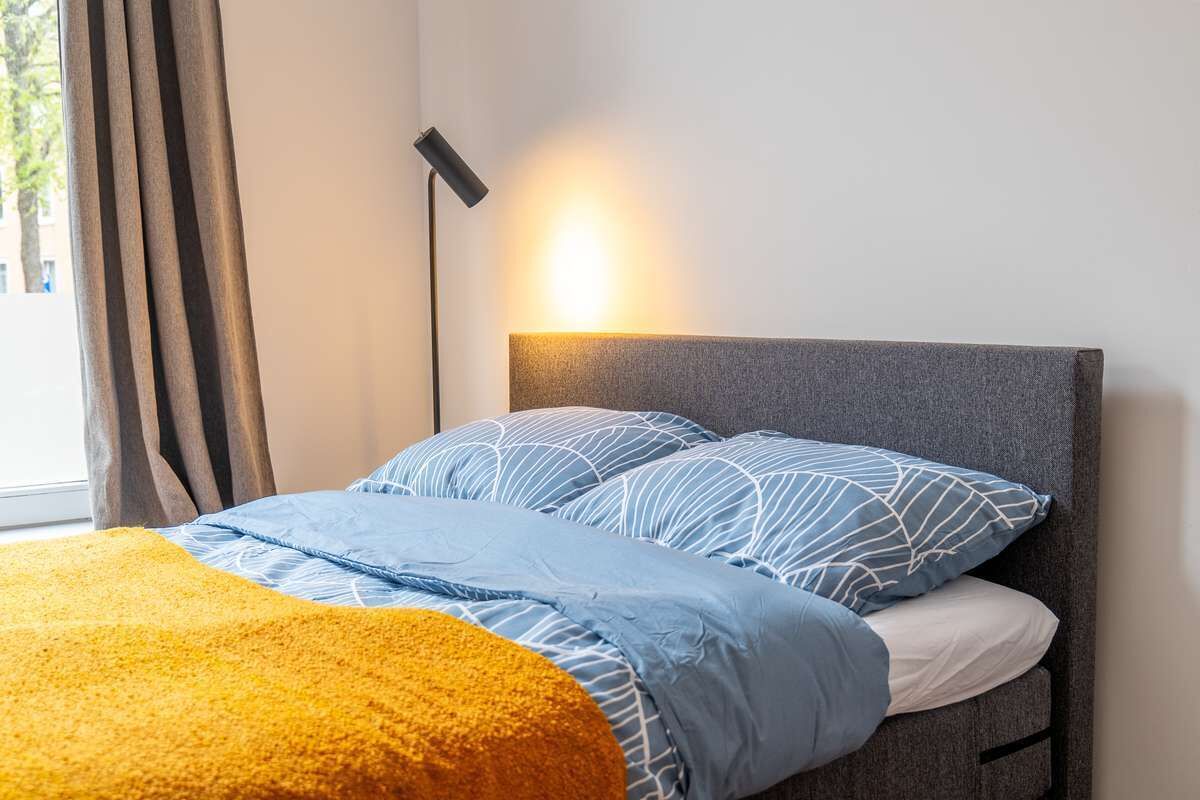 En seng med gråt sengegærde, blå og hvidstribet sengetøj og et gult tæppe, samt en gulvlampe.