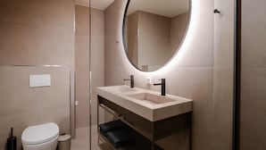 A modern bathroom with a double sink, a toilet, a trash bin, and a mirror with a circular light.