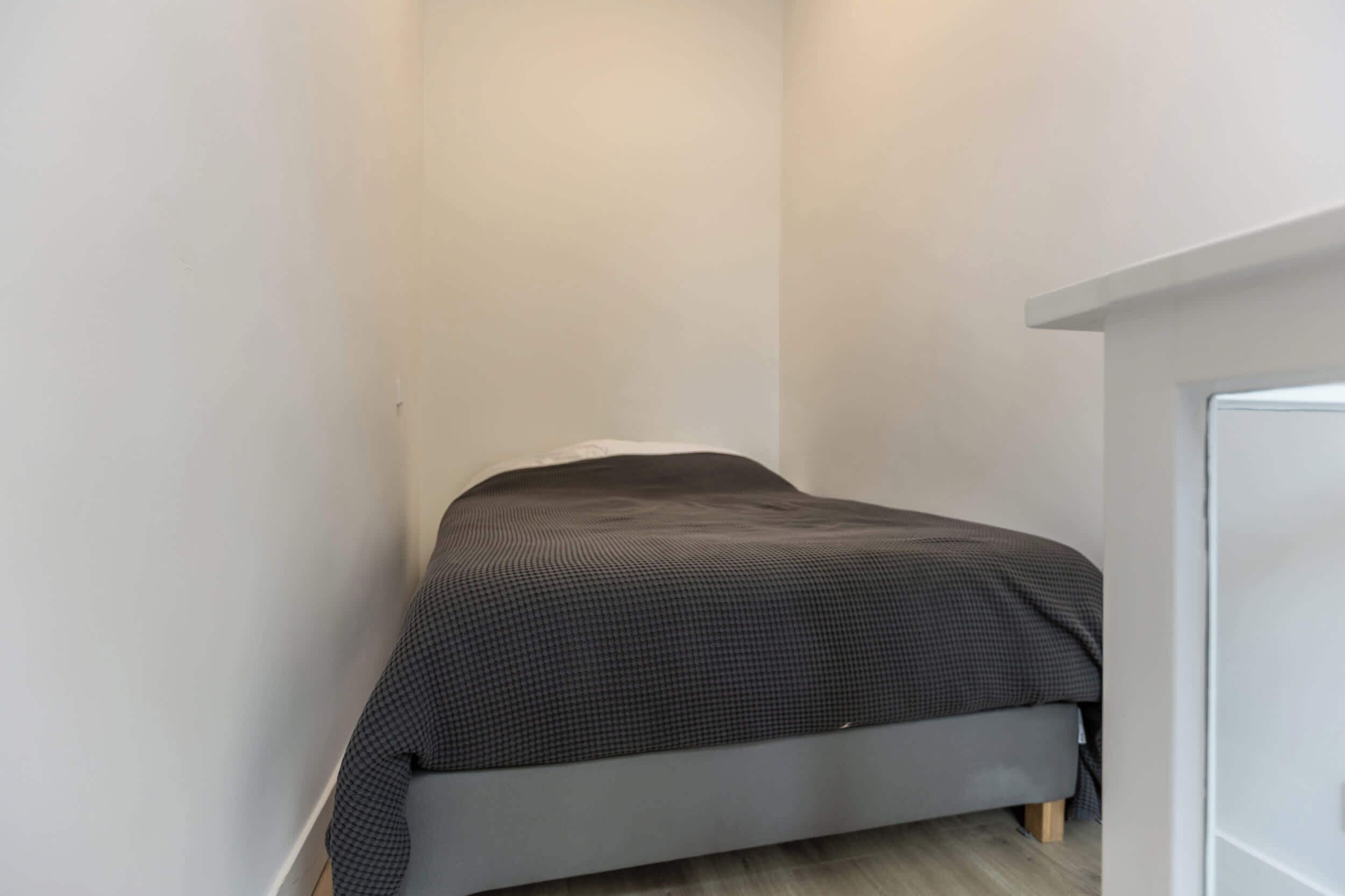 Une petite pièce étroite, avec un lit simple, des murs blancs et un plancher en bois.