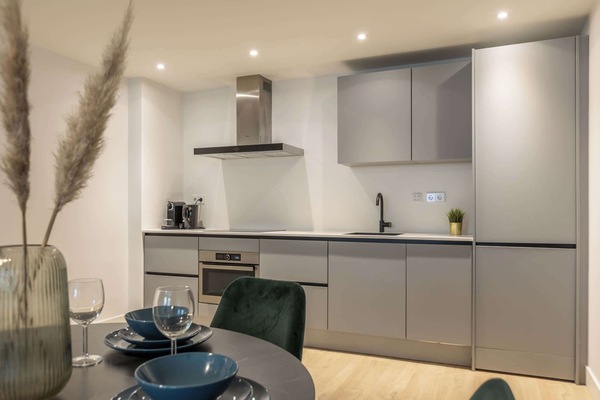 The fully equipped kitchen is integrated along the back wall and finished in matte grey cabinetry. It includes an oven, induction cooktop, extractor hood, fridge, and coffee machine.