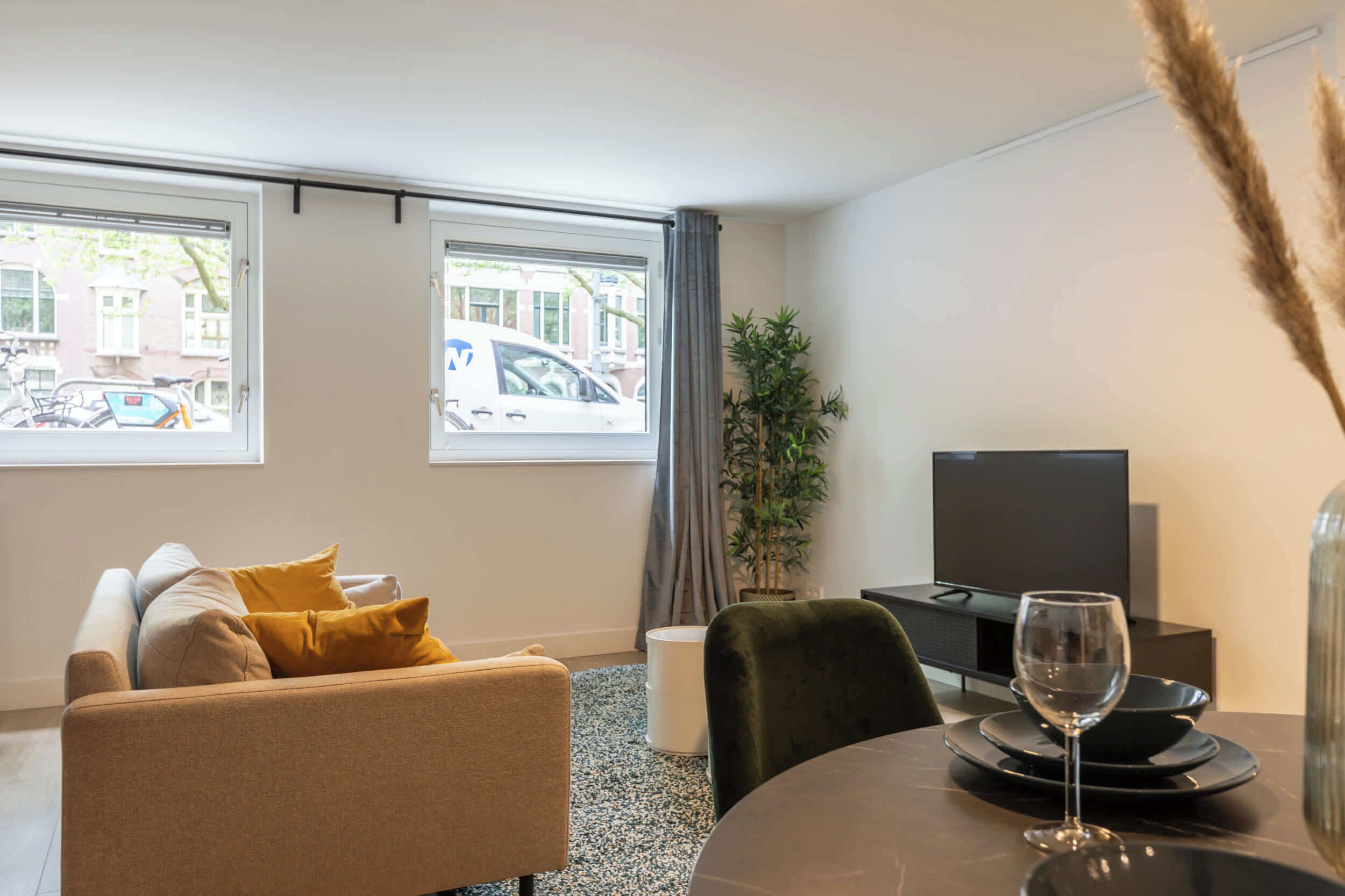 Una sala de estar moderna con un sofá, una mesa de comedor con una copa de vino, un televisor y ventanas amplias con vista a la ciudad.