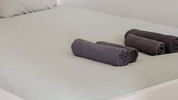Uma cama com colchão claro, um travesseiro e duas toalhas enroladas.