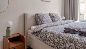 Un dormitorio con cama, mesita de noche, lámpara de pared, cuadro enmarcado y ventana con cortinas.