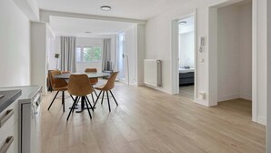 Eine moderne Küche mit Einbauofen, eine Essecke mit rundem Tisch und Stühlen sowie Blick in ein Schlafzimmer durch einen offenen Durchgang.