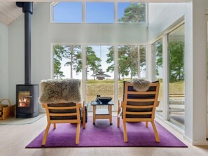 Ein Zimmer mit einem großen Fenster, einem Kamin, zwei gestreiften Sesseln, einem kleinen Tisch und einem violetten Teppich.