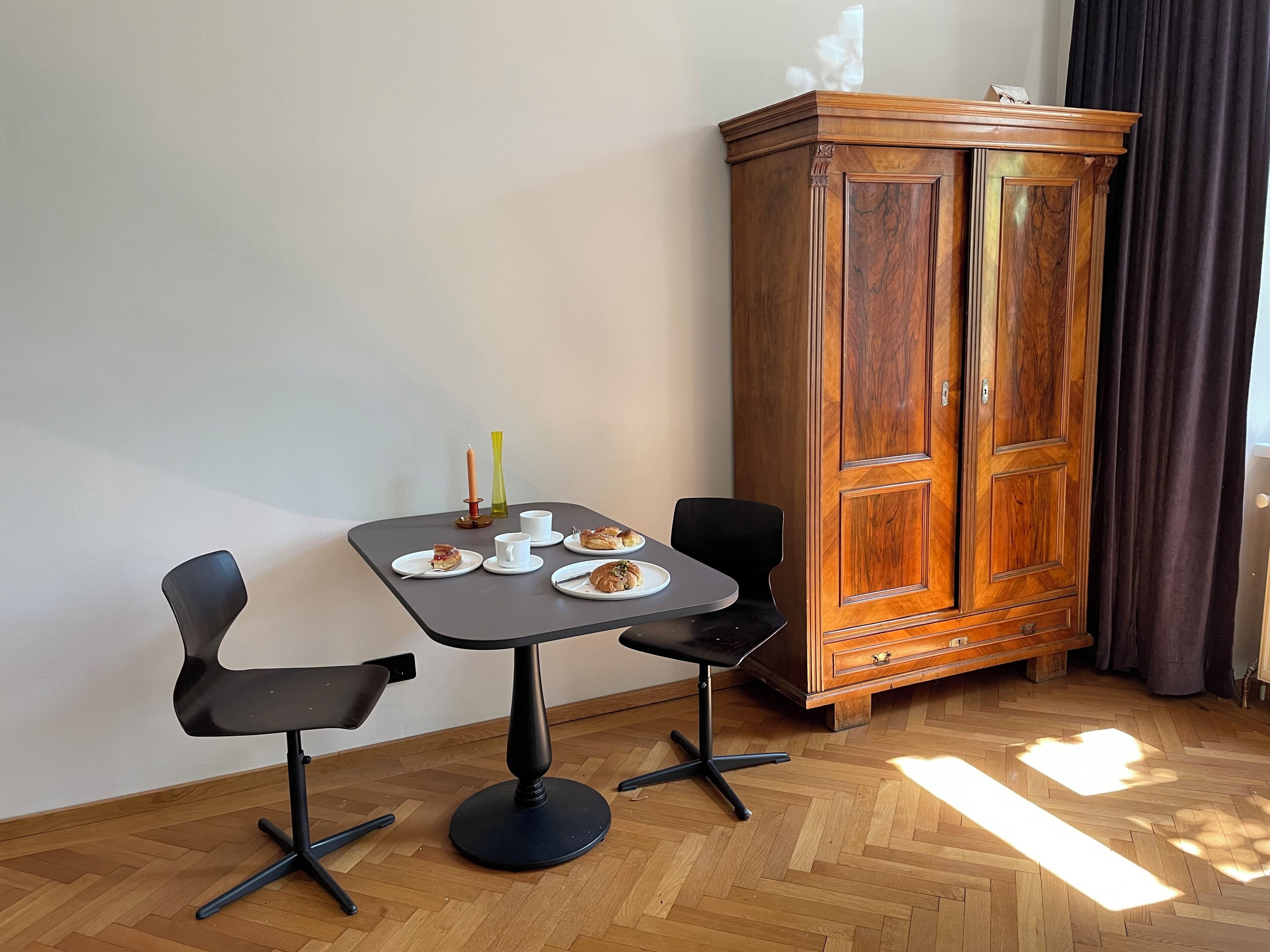 A room with a wooden cabinet, a small table with chairs, and a herringbone-patterned floor.