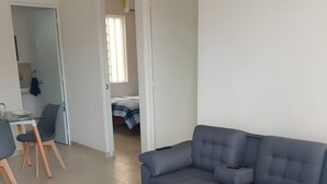 Un salon moderne avec un canapé d’angle gris, un petit tapis rond, un coin repas avec une table et des chaises, et une chambre à coucher visible au fond.