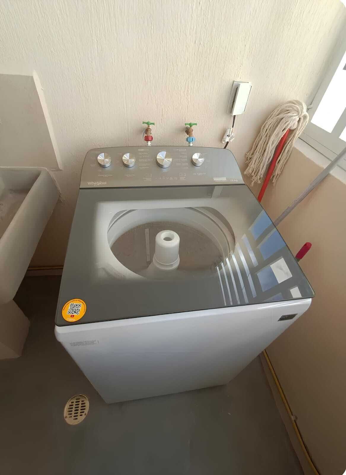 A compact, modern washing machine with a control panel, a small trash bin, and a mop on the wall.