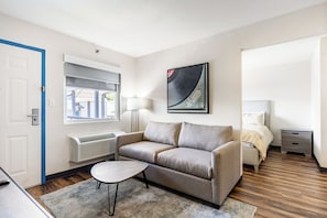 A modern hotel room with a sofa, a small table, a bed, a nightstand, a lamp, a radiator, a window, and a framed artwork on the wall.