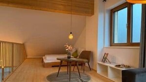 A cozy loft bedroom with a sloped ceiling, a round wooden table with a vase, a sofa, and a window with a view.