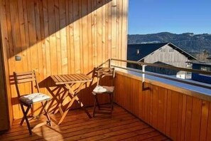 Eine Holzterrasse mit Tisch, StĂŒhlen, GelĂ€nder und Blick auf Berge und ein GebĂ€ude.