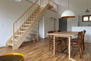 Ein gerĂ€umiges Zimmer mit einer Holztreppe, einem Esstisch mit StĂŒhlen und einer groĂen Pendelleuchte.