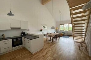 Eine moderne KĂŒche mit weiĂen SchrĂ€nken, einem Essbereich mit Holztisch und einem groĂen Fenster fĂŒr natĂŒrliches Licht.