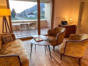 A room with a sofa, two armchairs, a small table, a lamp, and a view of a balcony with a table and chairs.