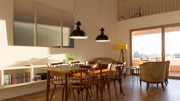 Un espace de salle à manger moderne avec une table et des chaises en bois, des luminaires suspendus, une bibliothèque et une vue dégagée sur l’extérieur grâce à de grandes fenêtres.