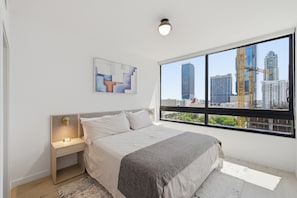 A bedroom with a bed, bedside table, nightstand, artwork, and a large window with a city view.