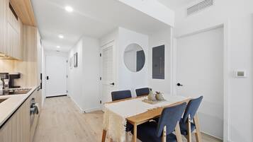 A modern kitchen with wooden cabinets and a built-in oven, a dining area with a wooden table and blue chairs, and a white hallway with a round mirror and a door.