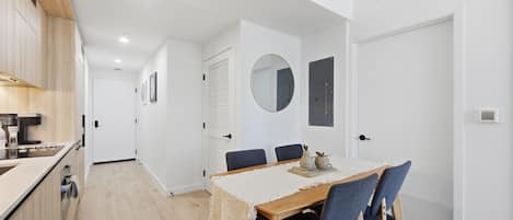 Une cuisine moderne avec des meubles en bois et un four encastré, un coin repas avec une table en bois et des chaises bleues, et un couloir blanc agrémenté d’un miroir rond et d’une porte.
