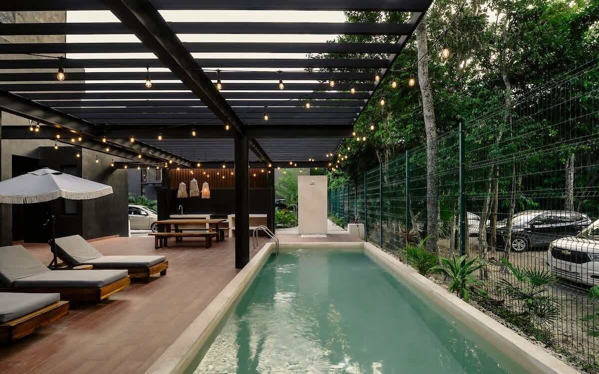 A pool area with lounge chairs, a pergola with string lights, and a fenced garden.
