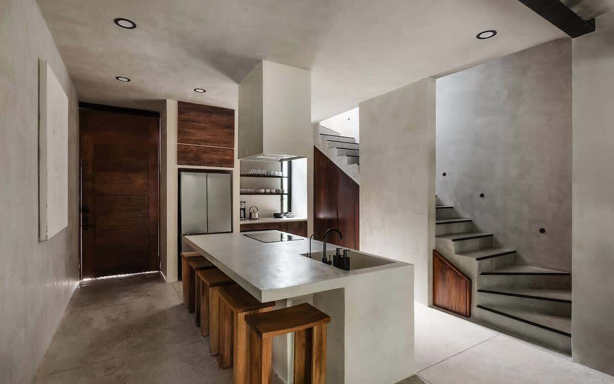 Une cuisine moderne avec un îlot central, des tabourets en bois et un escalier situé sur le côté droit.