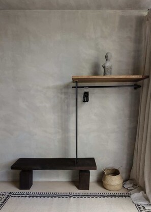 Ein schlichtes Zimmer mit einer dunklen Holzbank, einem einfachen Metallregal mit einer Skulptur und einem gewebten Korb auf dem Boden.