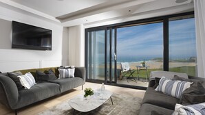 Ett modernt vardagsrum med en grå soffa, ett soffbord och en stor skjutdörr i glas som leder ut till en balkong med utsikt över stranden.