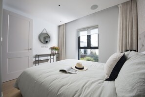 Ein Schlafzimmer mit Bett, Nachttisch mit Blumenvase, Fenster mit Vorhängen und einer Tür.