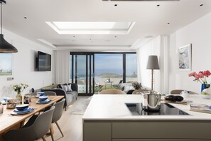 Ein modernes Wohnzimmer mit großem Fenster und Meerblick, ein Essbereich mit gedecktem Tisch und eine Küche mit Kochinsel.