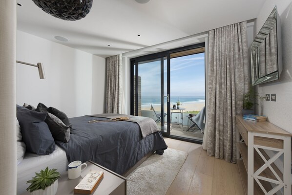 Ein Schlafzimmer mit einem Bett, einem Nachttisch mit Buch und Tasse, einer kleinen Pflanze und Blick auf den Strand durch eine Schiebetür.
