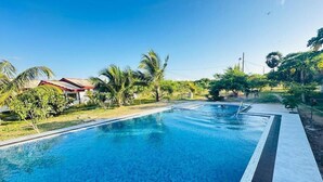 Una piscina con agua azul cristalina, rodeada de vegetación y algunos edificios al fondo.
