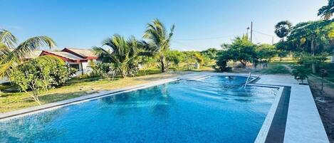 Una piscina con agua azul cristalina, rodeada de vegetación y algunos edificios al fondo.