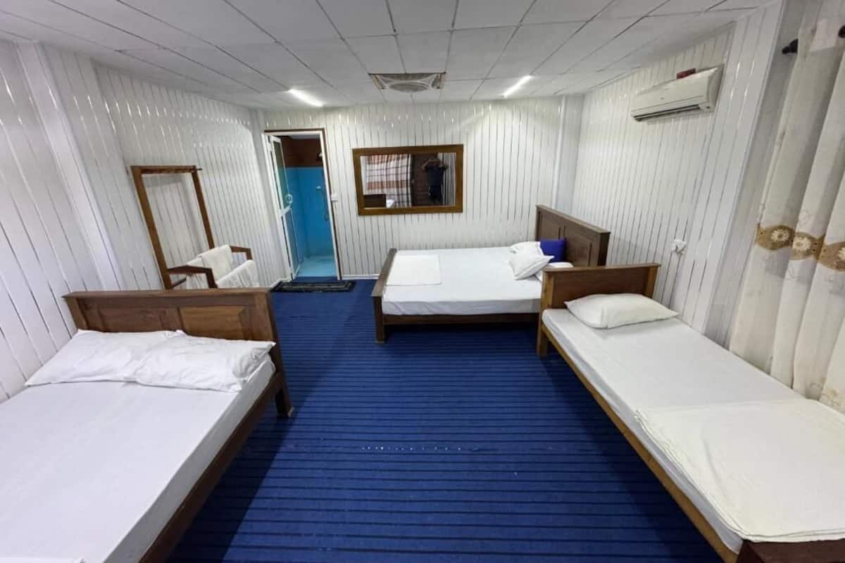 Habitación de hotel con tres camas individuales, alfombra azul, paredes blancas y cabecero de madera.