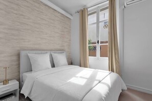 Ein Schlafzimmer mit einem Bett, einem Nachttisch, einem Fenster mit Vorhängen und Blick ins Freie.
