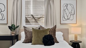 A neatly arranged bedroom with a bed, pillows, a bedside table, a lamp, and a window with blinds. - Spacious 7BD/7BR Home in Somerville, 10 mins to Boston, Harvard, MIT (Somerville)