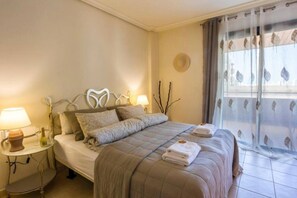 Ein Hotelzimmer mit Bett, Nachttischlampen, einem kleinen Tisch und Blick ins Freie durch transparente Vorhänge.