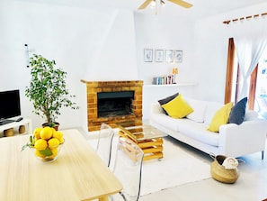 A living room with a fireplace, a white sofa with yellow cushions, a glass coffee table, and a potted plant.