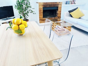 A modern living room with a wooden dining table, a glass bowl of lemons, a brick fireplace, a white sofa, and a TV.