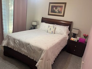 Una cama bien hecha con una colcha estampada con motivos florales, un cabecero de madera, una mesita de noche con una lámpara, un cuadro en la pared y una pequeña mesa con un bolso rosa y un letrero que dice 'HAPPY'.