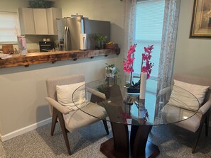 A dining area with a glass table surrounded by two chairs, a kitchen with a refrigerator and cabinets, and a window with curtains. - Casa familiar cerca de Stockyards (Fort Worth)